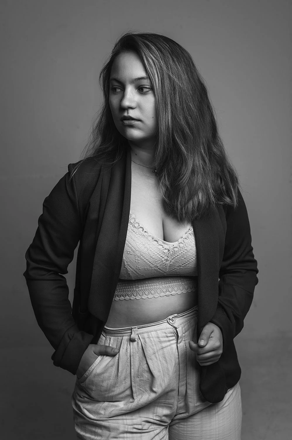 Frau mit einem dunklen Blazer, einem hellen Top und einer hellen Hose steht vor einer hellen Wand. Sie hat eine Hand lässig in der Hosentasche und ihr Blick geht verträumt nach Links.